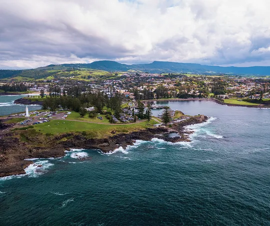Aerial view of a coastal town with a lighthouse, rocky shoreline, green hills, and cloudy sky.