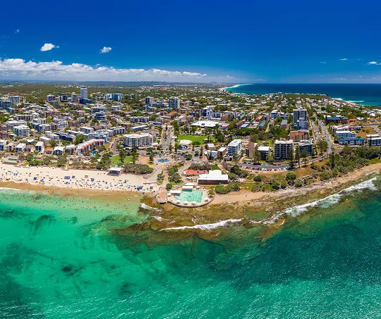 Aerial view of a coastal city with turquoise ocean, sandy beach, and urban buildings under clear blue sky.