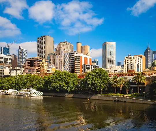 City skyline with modern buildings and green trees along a riverbank under a blue sky.