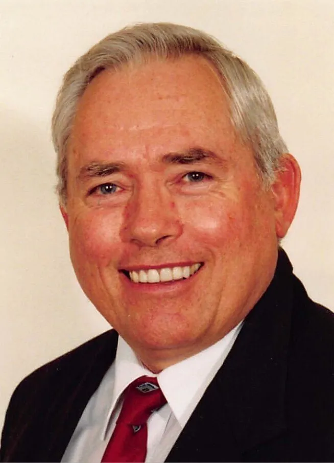 Portrait of an older man with gray hair wearing a black suit, white shirt, and red tie.