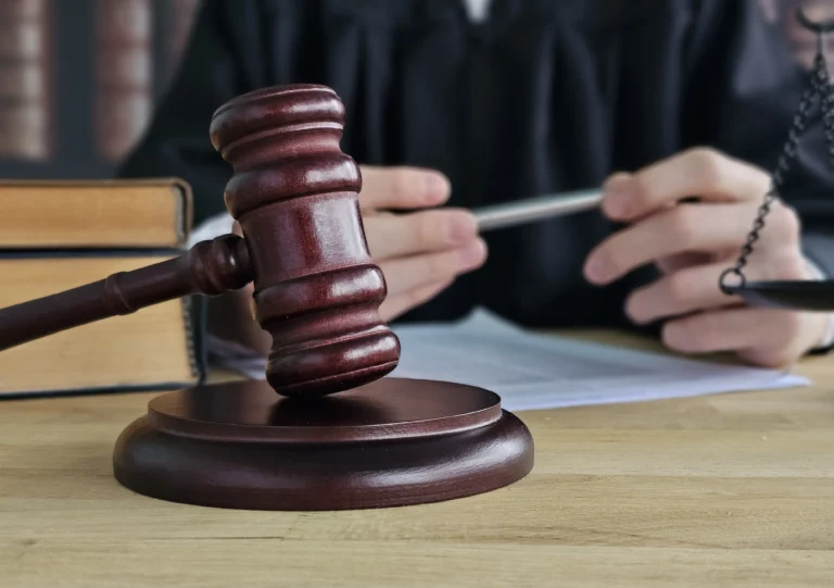 Close-up of a wooden judge's gavel on a desk with legal books and a person in judicial robes holding a pen.